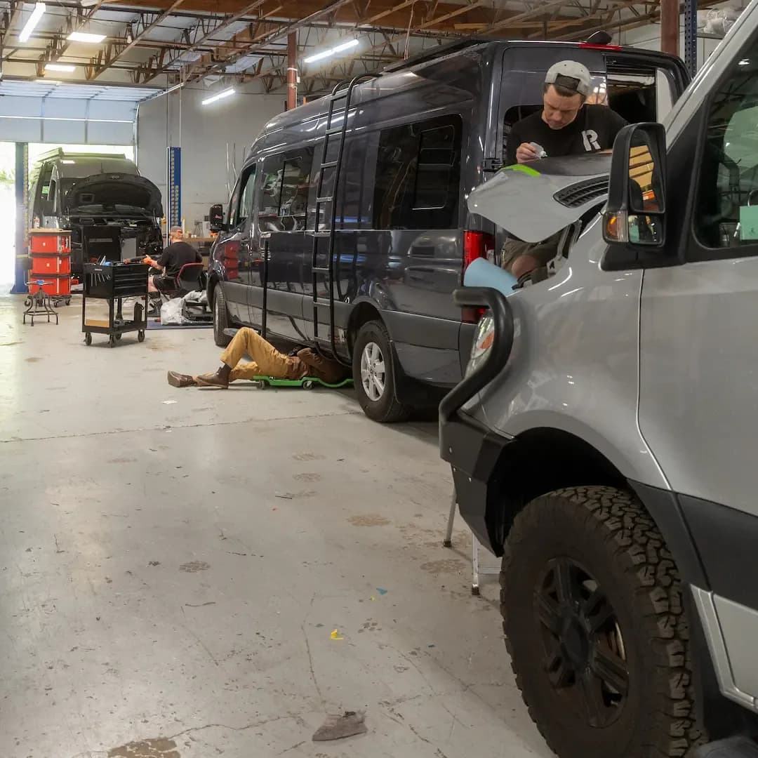 The Reparadise shop floor with several vans in service, one technician working a hood and another on a creeper underneath