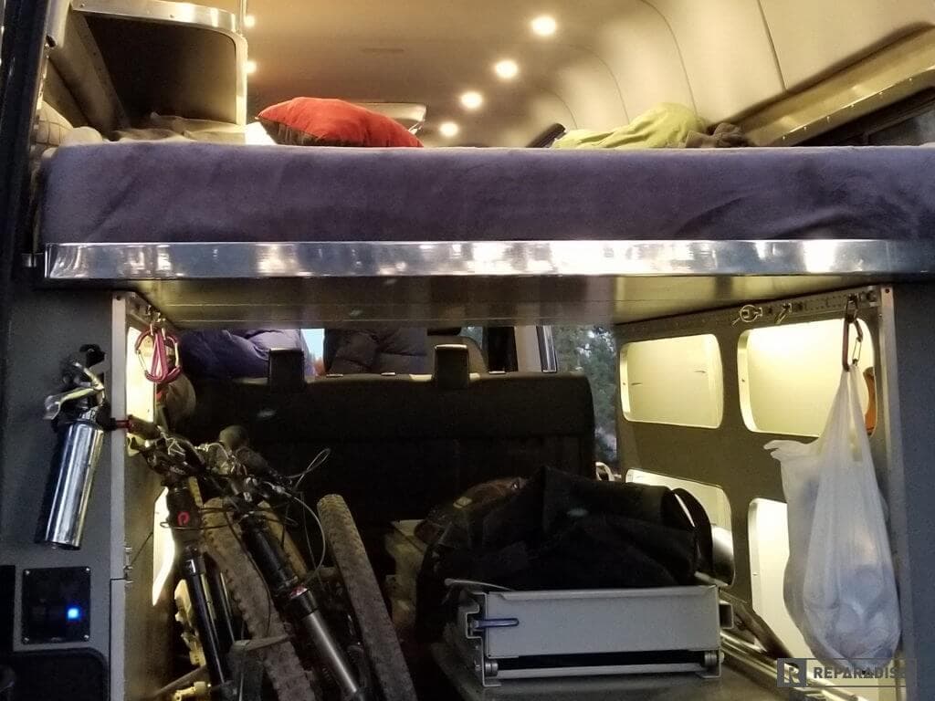 Fold-away upper sleeping platform with mattress, under-bed bike storage, and overhead lighting in 4x4 Sprinter Camper Van