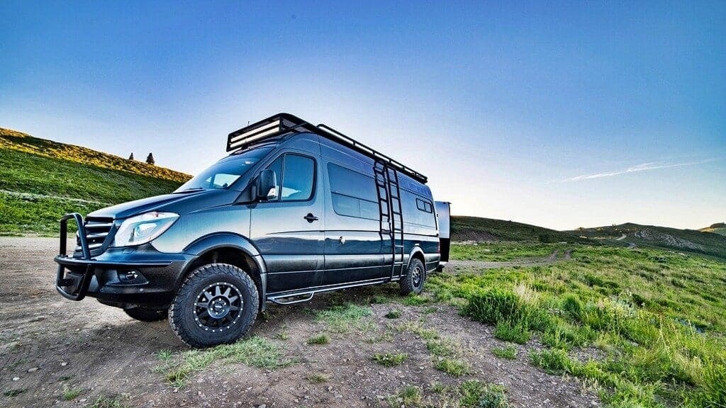 Black Mercedes Sprinter van with custom roof rack, ladder, front bull bar, and off-road wheels in wilderness setting 4x4 Sprinter Camper Van